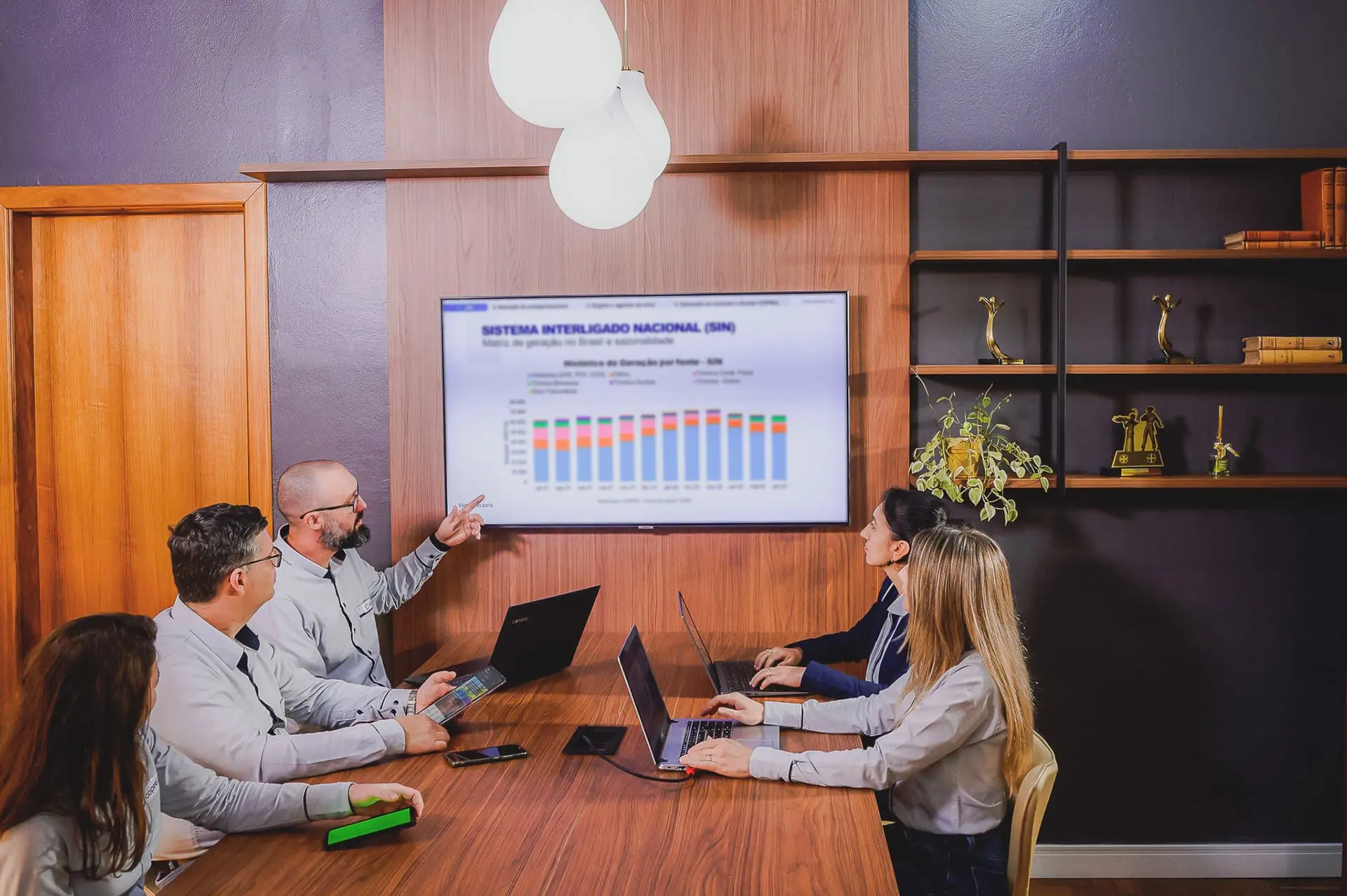 Grupo de pessoas em sala de reuniões analisando resultados em um gráfico exposto em uma televisão